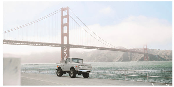 A white vintage pickup truck drives along a waterfront road with the Golden Gate Bridge and water in the background under a hazy sky.
